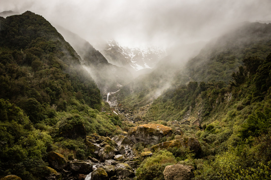 A steep mountain river flowing from misty snowcapped peaks into a lush valley
